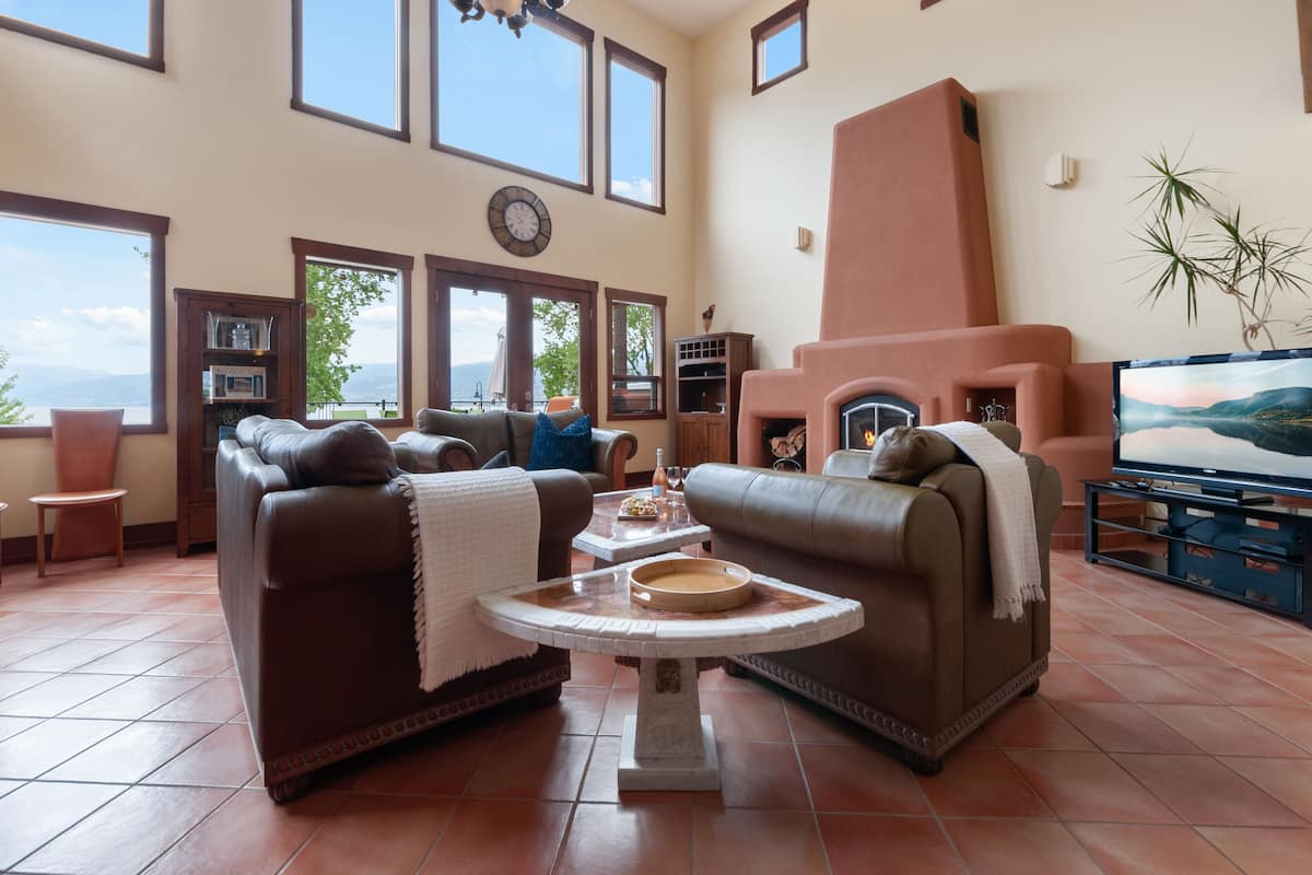 Un salón amplio con sofás de cuero, chimenea, televisor y ventanas grandes con vistas al exterior.
