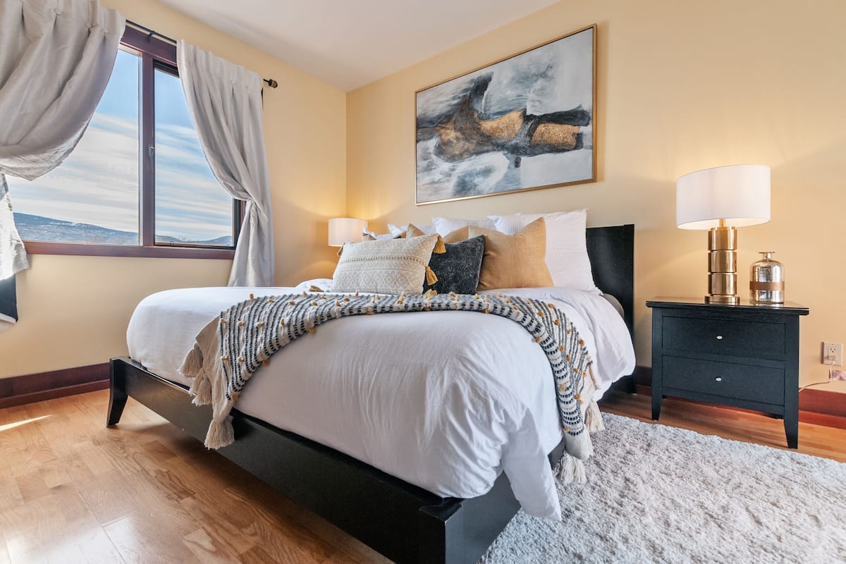 Un dormitorio con cama, mesitas de noche, ventana con cortinas, un cuadro en la pared y una lámpara.