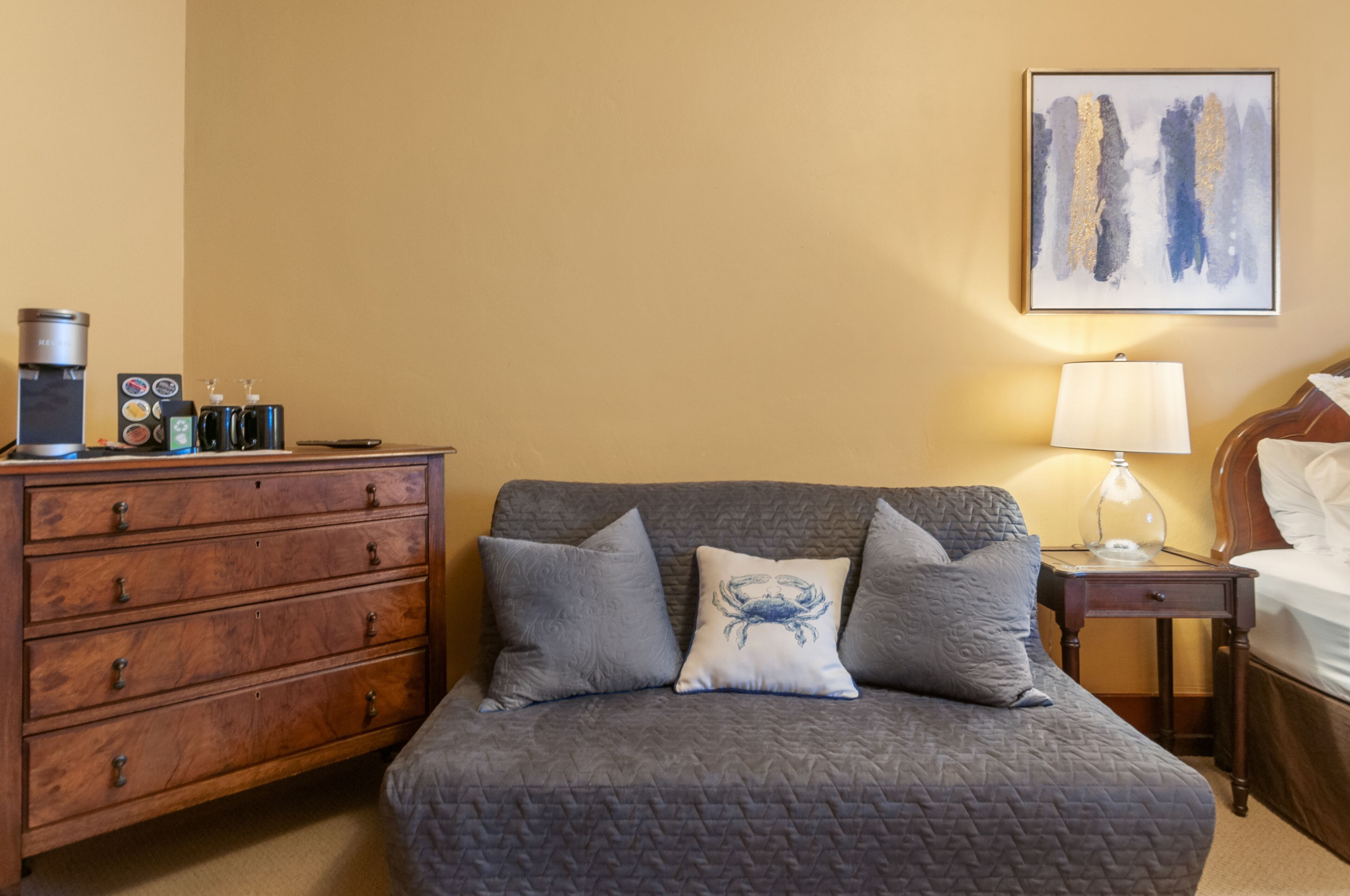 Habitación de hotel con sofá gris acolchado, cómoda de madera, mesita de noche con lámpara y un cuadro abstracto enmarcado en la pared.