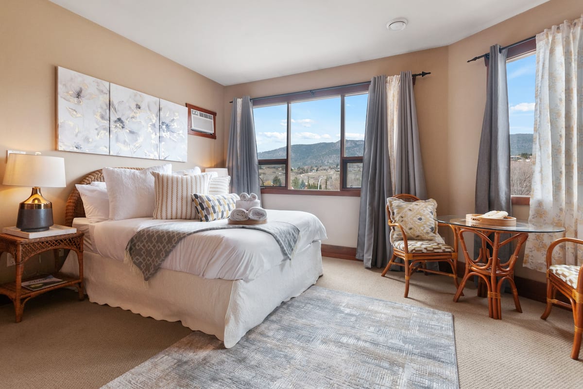 Un dormitorio con una cama grande, una mesita de noche con lámpara, una silla, una pequeña mesa y vistas a las montañas desde la ventana.