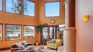 A spacious living room with large windows, a chandelier, a clock, and a view of the cityscape. - Casa Grande Inn (Penticton)