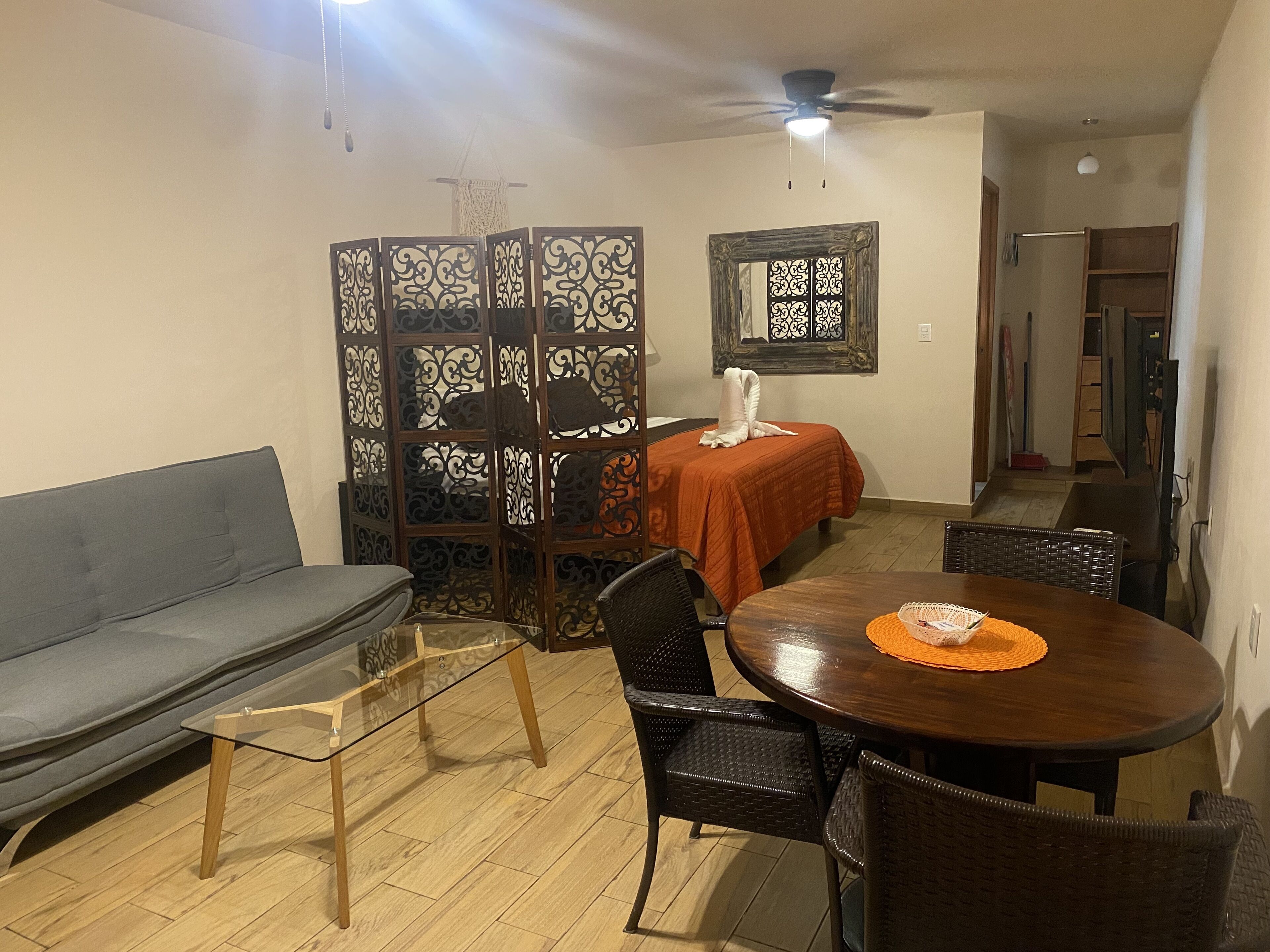 A compact living space with a sofa, a glass-top coffee table, a dining table with chairs, a decorative partition, and a ceiling fan.
