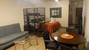 A compact living space with a sofa, a glass-top coffee table, a dining table with chairs, a decorative partition, and a ceiling fan.