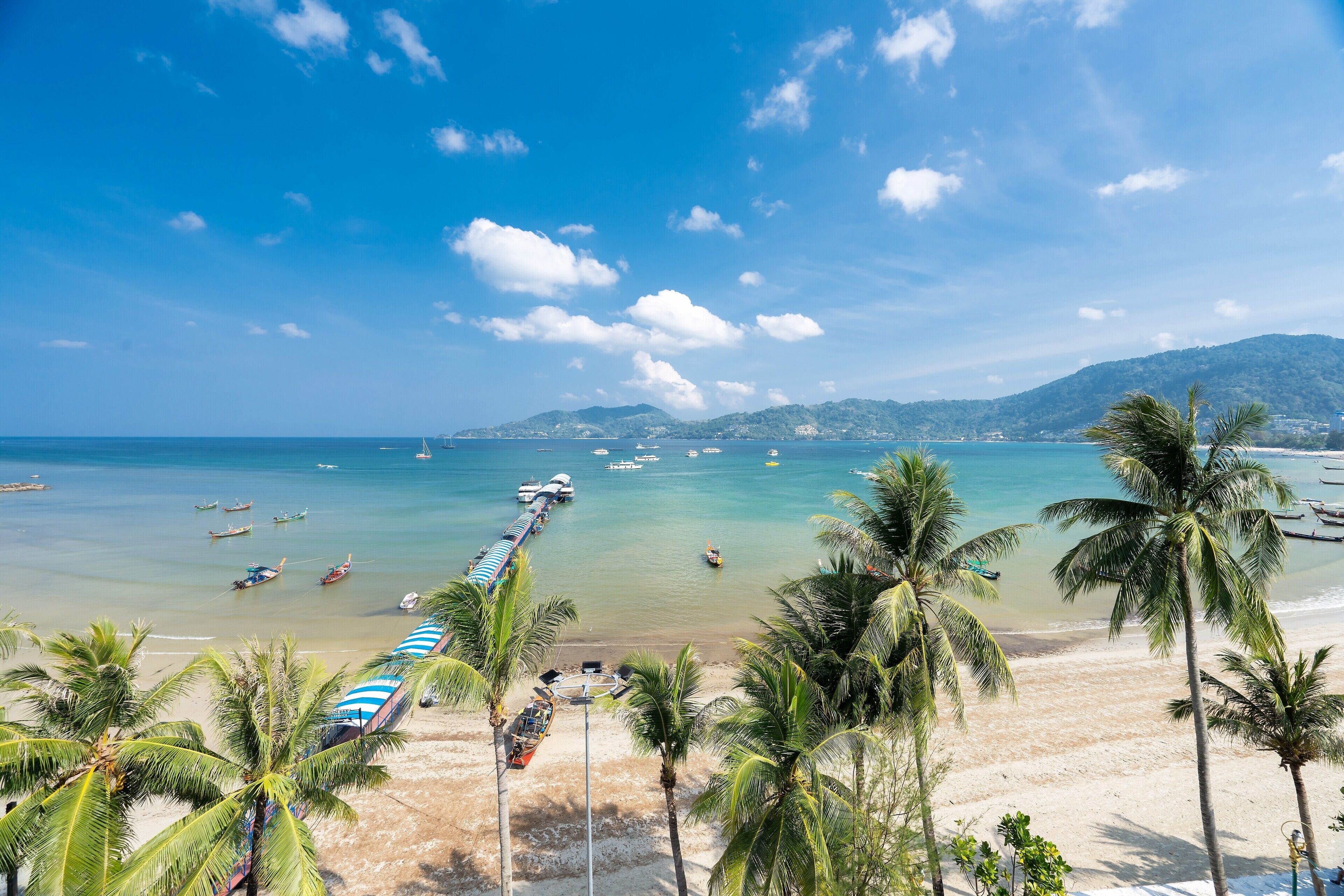 A beach with palm trees, a pier, and boats. - Patong Signature Boutique Hotel (Patong)
