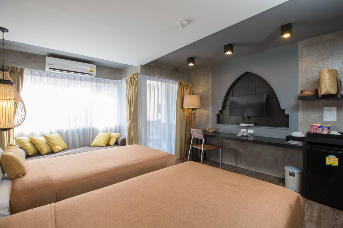 Ein Hotelzimmer mit zwei Betten, einem Schreibtisch, einem Stuhl, einer Klimaanlage, einem Fernseher und einer kleinen Küchenzeile.