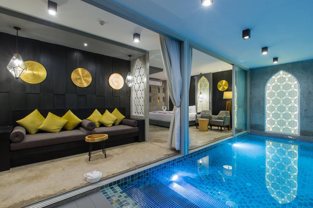 Ein moderner Poolbereich im Innenbereich mit einer Couch und einem kleinen Tisch, einem großen Spiegel, der das Schwimmbecken reflektiert, und einer Wand mit Muster.