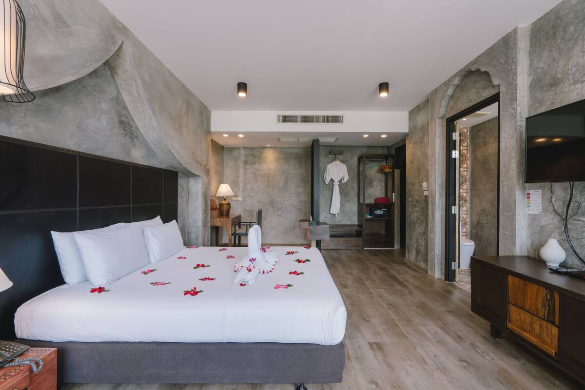 Ein modernes Hotelzimmer mit einem großen Bett, einem Holz-Kopfstück, einem Flachbildfernseher, einer Holzkommode und einem durch eine offene Tür einsehbaren Badezimmer.