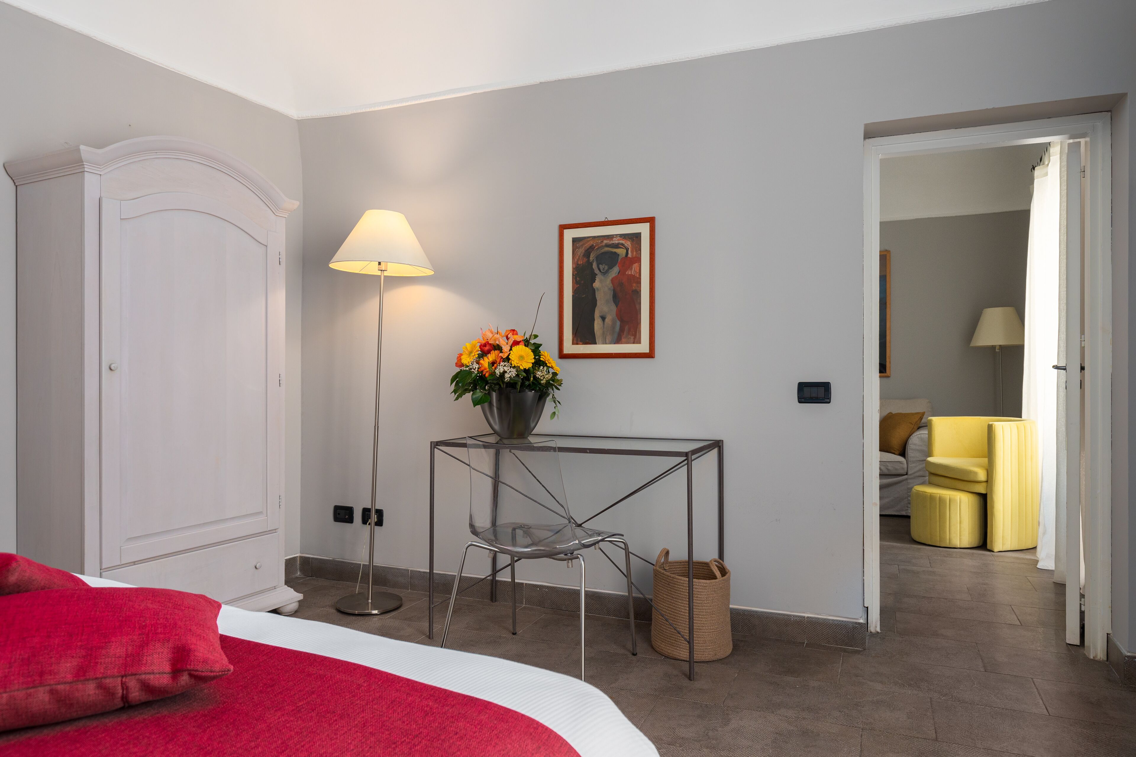 Habitación de hotel con cama, escritorio con lámpara, silla, un jarrón de flores y un cuadro en la pared.