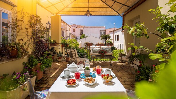 Un área de comedor al aire libre iluminada por el sol, con una mesa dispuesta para el desayuno, rodeada de plantas en macetas y vista a la calle.