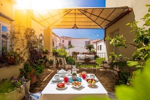 Un área de comedor al aire libre iluminada por el sol, con una mesa dispuesta para el desayuno, rodeada de plantas en macetas y vista a la calle.