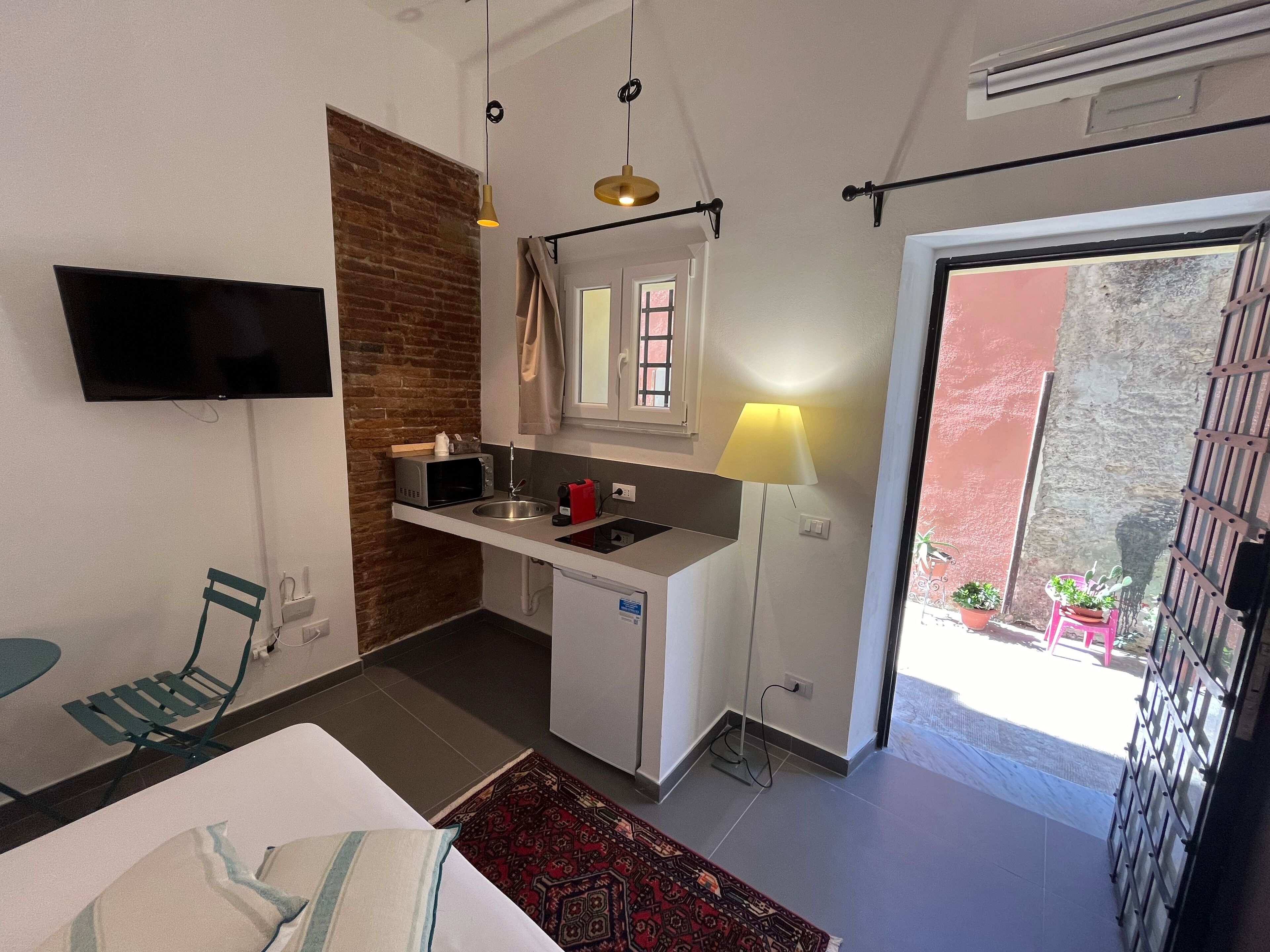 A compact kitchen with a microwave, sink, and a small refrigerator. There is a TV mounted on the wall, a chair, and a table with a patterned rug. A balcony with potted plants is visible through an open door.