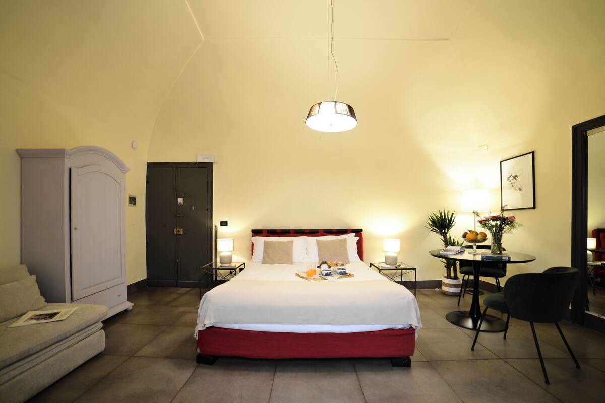 A hotel room with a bed, bedside tables, a desk with a chair, a wardrobe, and a door.