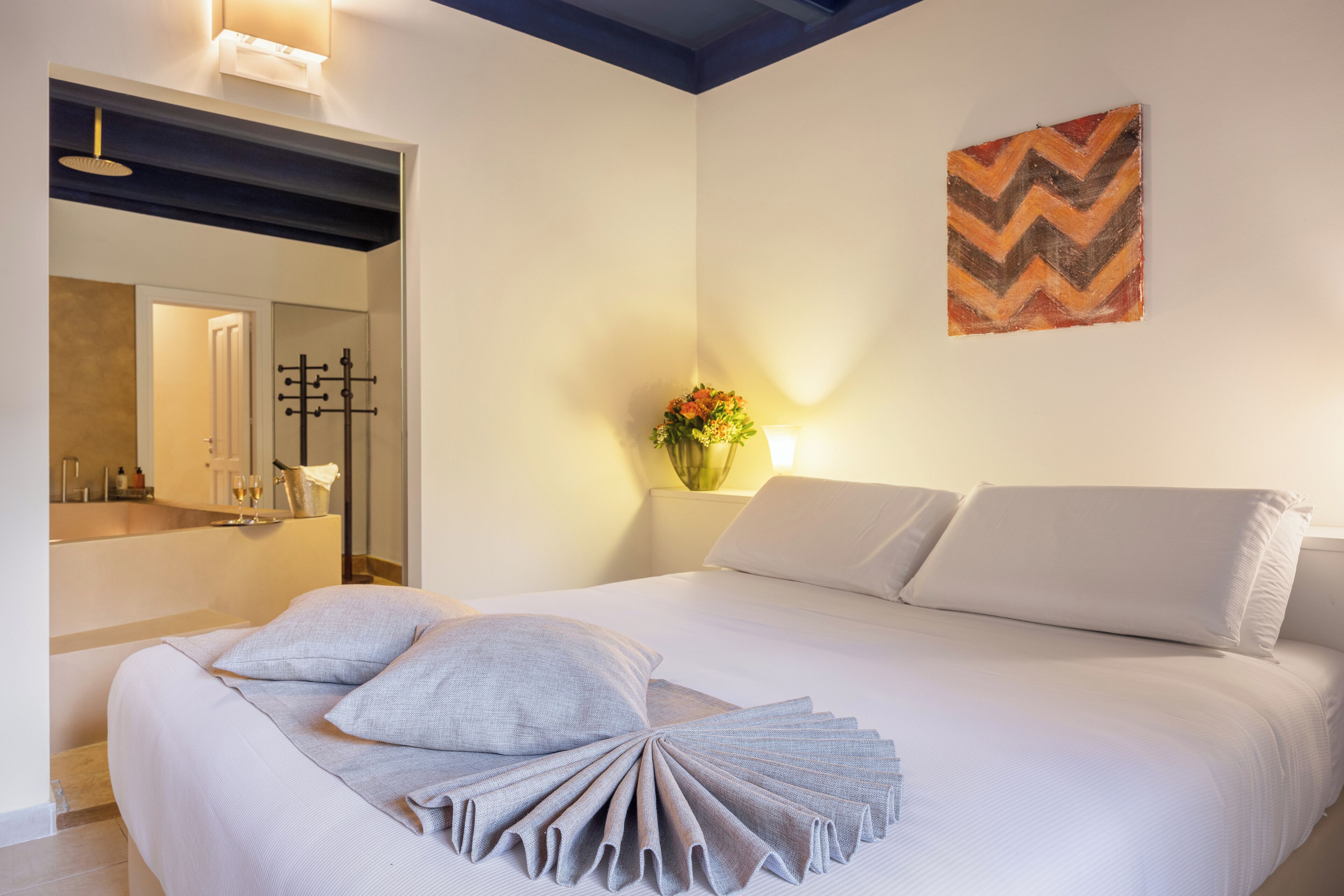 A modern hotel room with a large bed, a mirror, a decorative wall piece, and a vase with flowers.