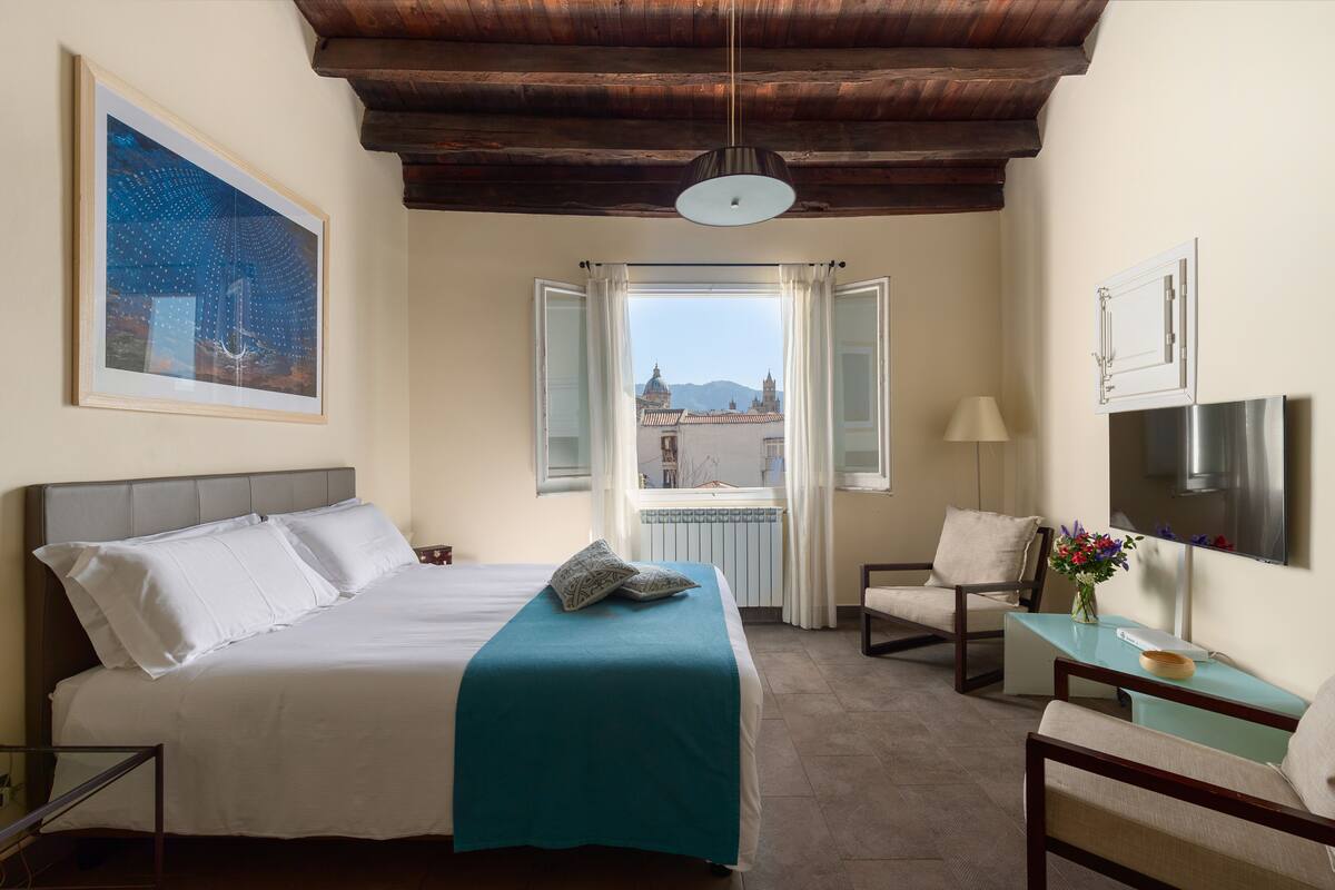 A hotel room with a bed, a desk, a chair, a window with curtains, a painting on the wall, and a view of buildings outside.