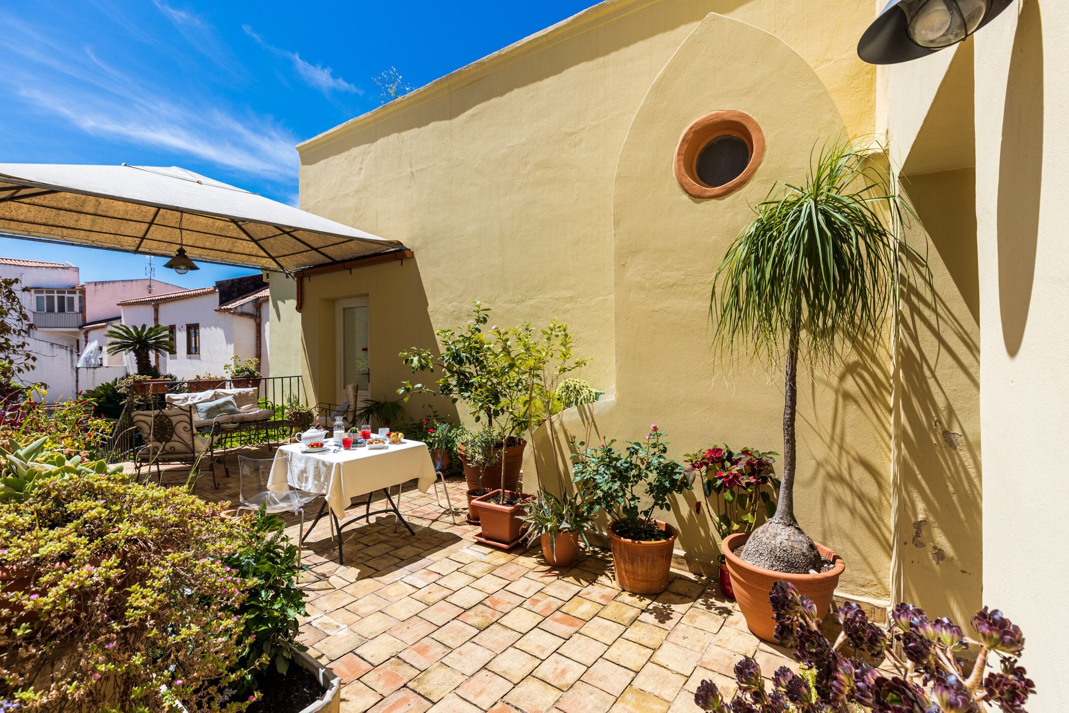 A patio with a table set for dining, surrounded by potted plants and a sun umbrella.