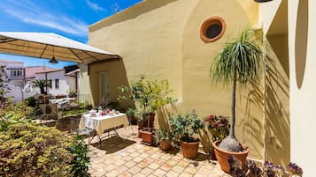 Un patio con una mesa lista para comer, rodeado de macetas con plantas y una sombrilla.