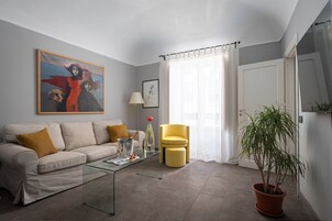 A living room with a sofa, a glass coffee table, a yellow armchair, a potted plant, and a painting on the wall.