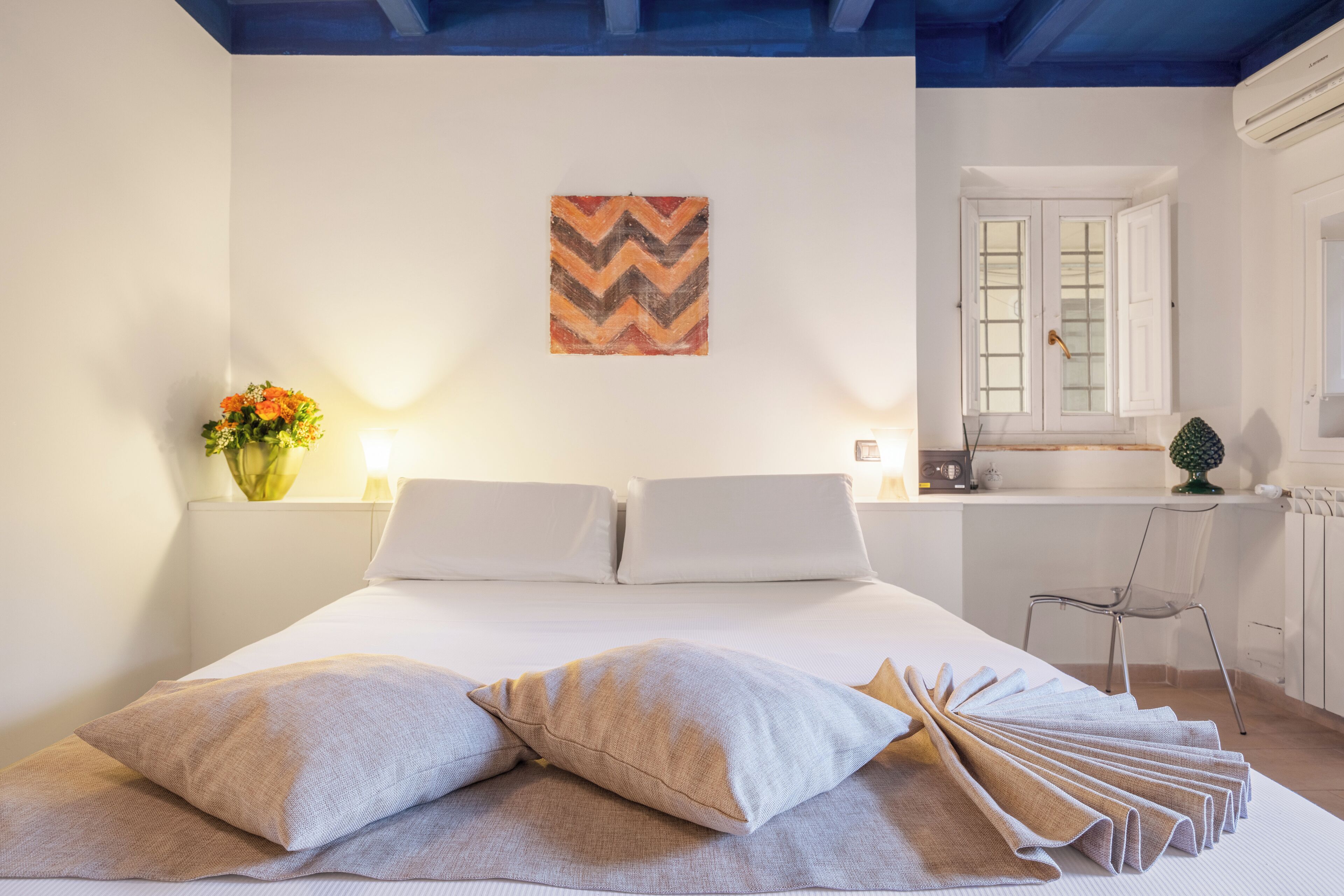 Uma cama bem arrumada com travesseiros e uma toalha dobrada, uma obra de arte na parede, uma janela com cortinas e uma cadeira em um quarto claro com paredes brancas e vigas azuis no teto.