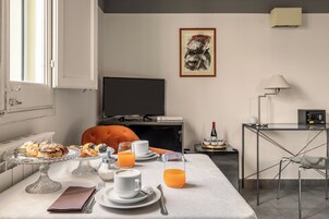 Desayuno con pasteles, café y zumo de naranja sobre una mesa, un televisor y un escritorio con lámpara y radio en una habitación con ventana y un cuadro en la pared.