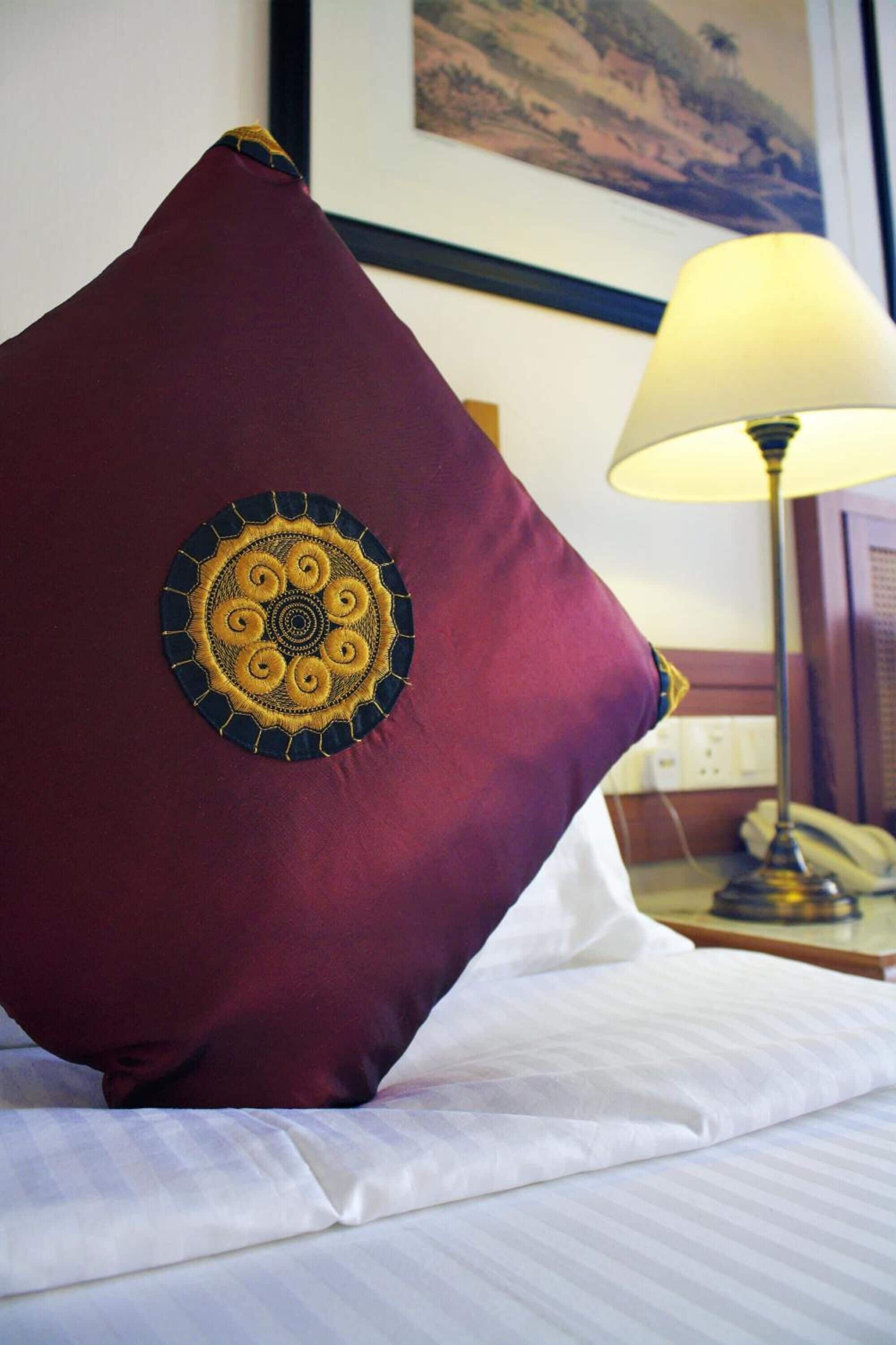 En seng med en bordeaux pude med et cirkulært broderet motiv, en hvid pude og en lampe på et natbord.