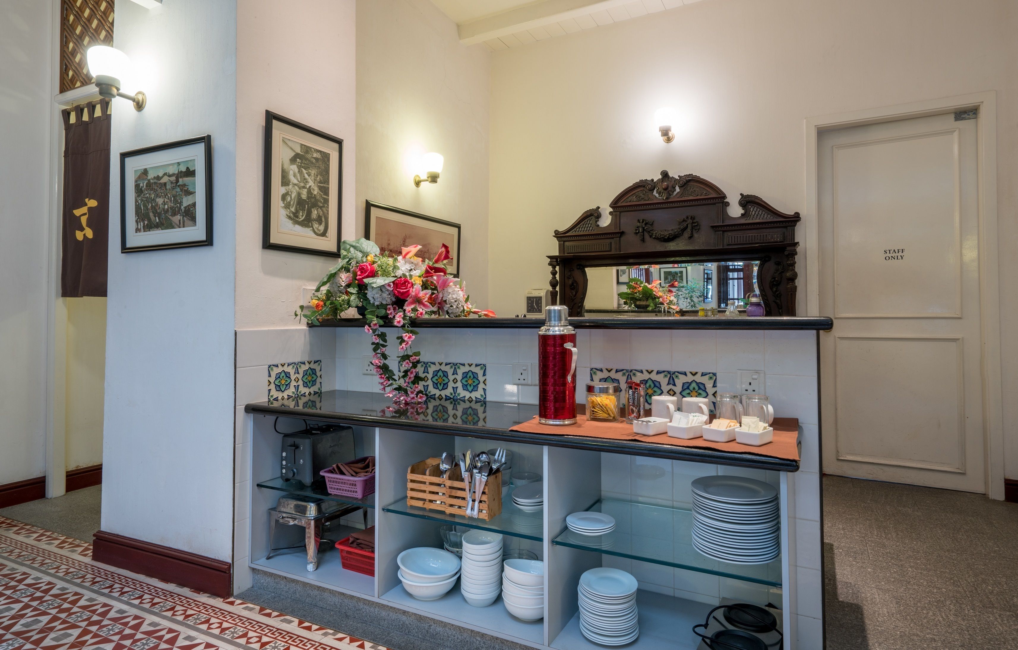 A reception area with a counter displaying plates, bowls, and a vase of flowers. There are framed pictures on the wall, a mirror above the counter, and a 'STAFF ONLY' door to the right.