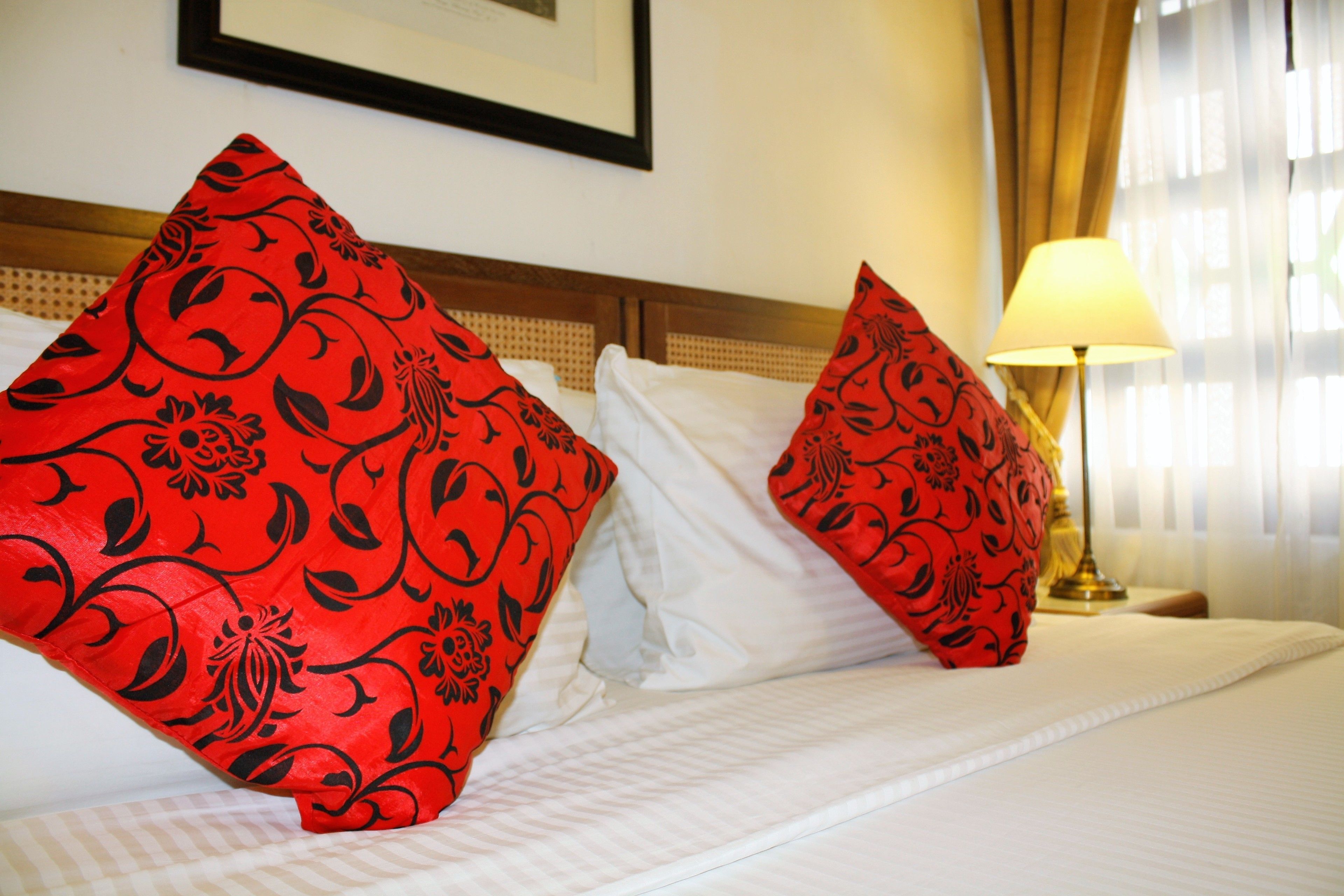 A hotel room with a bed, two red patterned pillows, a white pillow, a lamp, and a framed picture on the wall.
