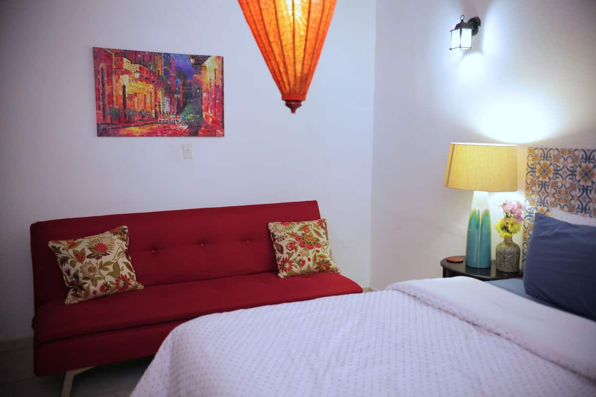 A room with a red sofa, a bed with a white bedspread, a painting on the wall, a table lamp, and a decorative vase.