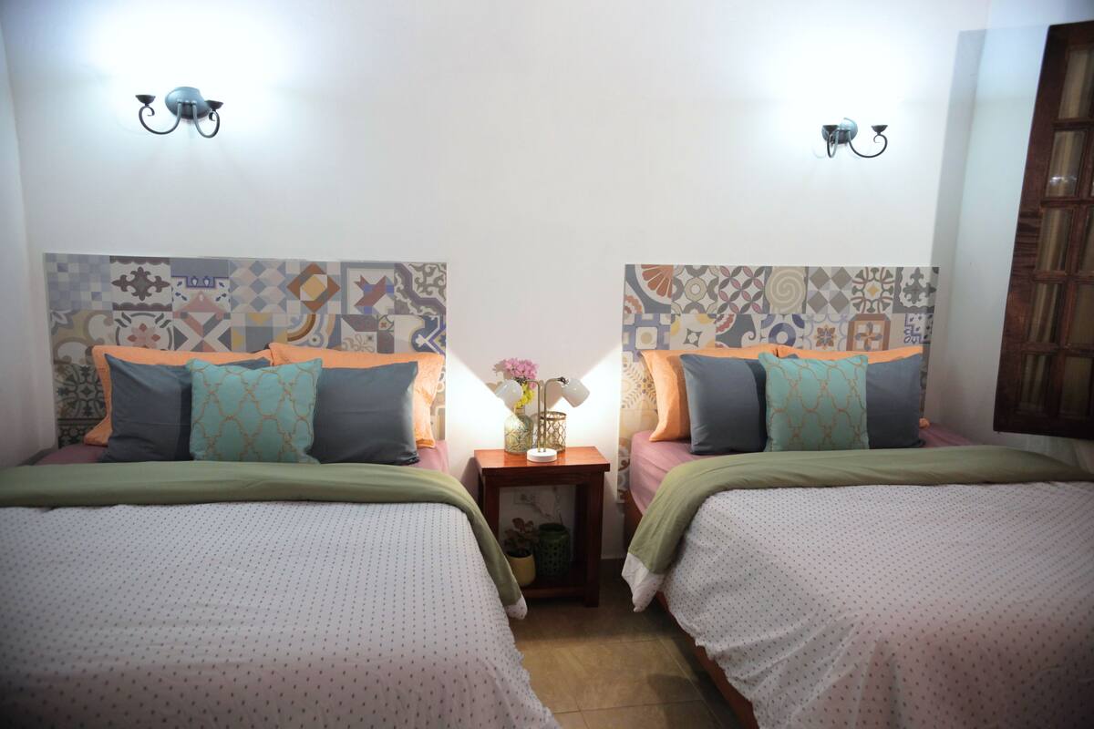 A room with two beds, each with patterned headboards and matching pillows. There is a small table with a lamp and a vase with flowers between the beds.