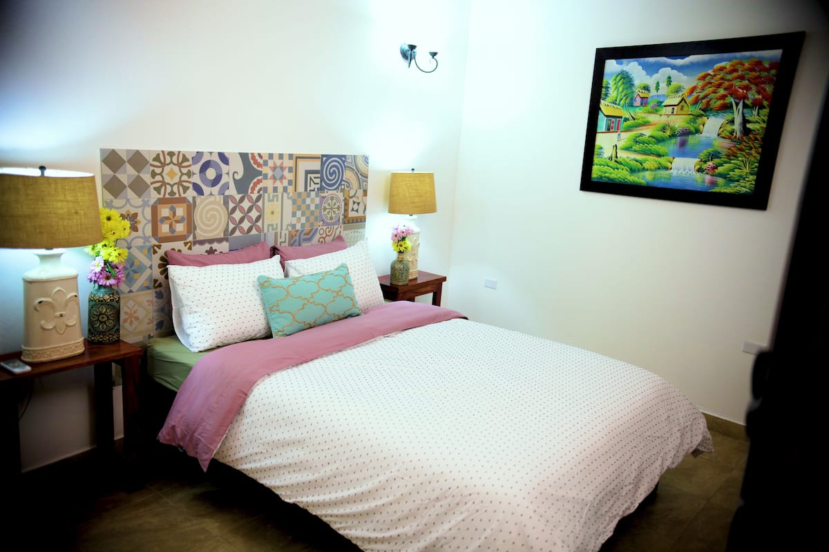 A neatly arranged bedroom with a patterned headboard, a bed with a pink blanket, two bedside tables with lamps and decorative items, and a wall-mounted painting.