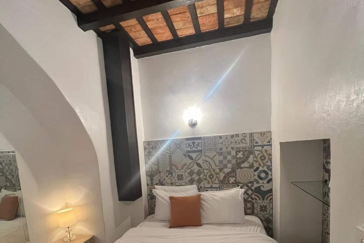 A bedroom with a bed, a nightstand, a wall-mounted light, and a patterned tile wall.
