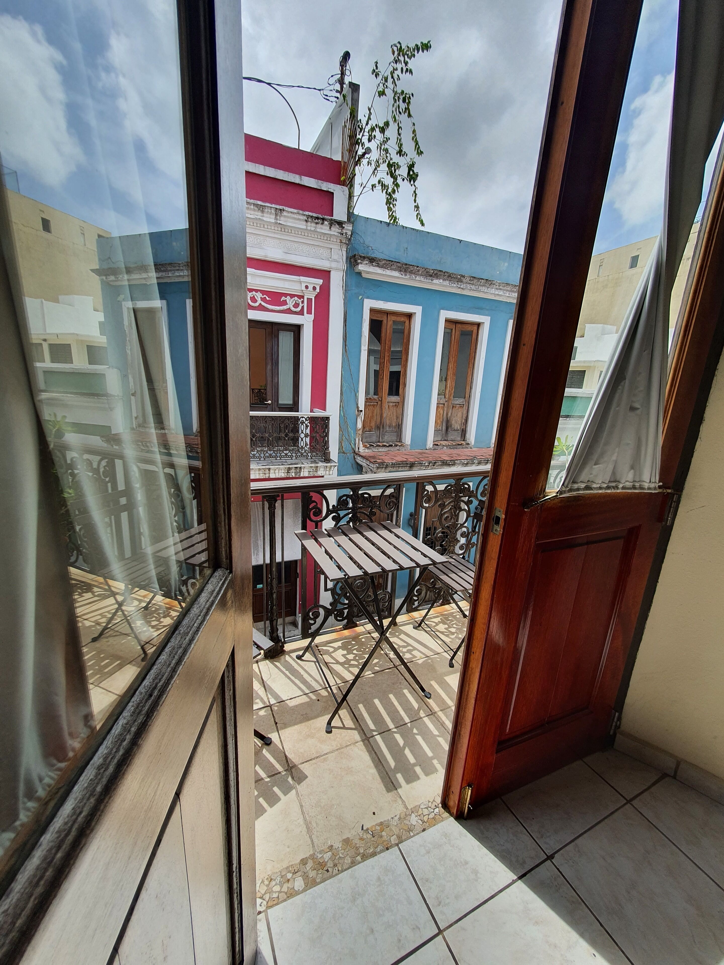 A balcony with a wooden table and chairs, a view of colorful buildings, and a tiled floor.