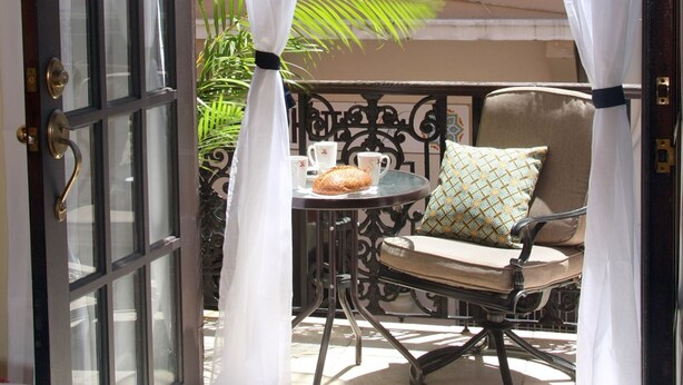 An open door with white curtains, a chair with a cushion, a small table with a cup and saucer, and a potted plant. - Fortaleza Suites (San Juan)