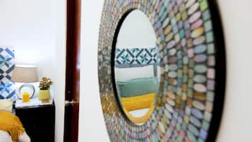Espejo con marco de mosaicos que refleja una pared con diseño, y una pequeña mesa con una lámpara, un florero y una vela.