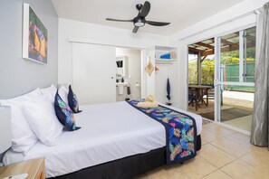 A hotel room with a bed, a ceiling fan, a bathroom with a sink and mirror, and a sliding glass door to an outdoor area.
