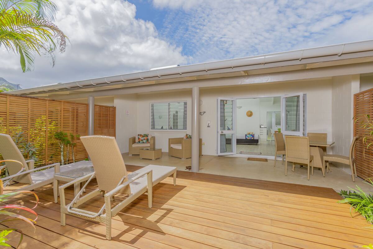 A covered outdoor patio with wicker furniture, a wooden deck, and a view of the interior through glass doors.