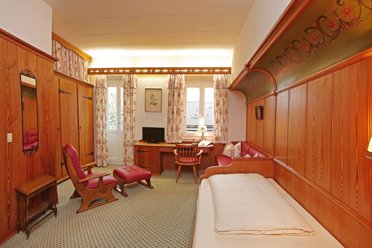 A hotel room with a wooden wardrobe, a desk with a chair, a bed, and a television.