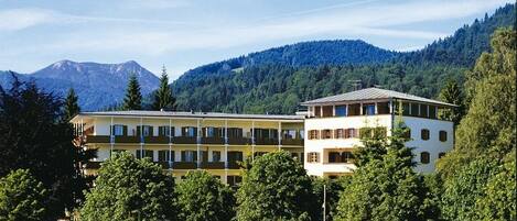 Un hotel de varios pisos con techo plano, rodeado de exuberante vegetación y montañas al fondo.