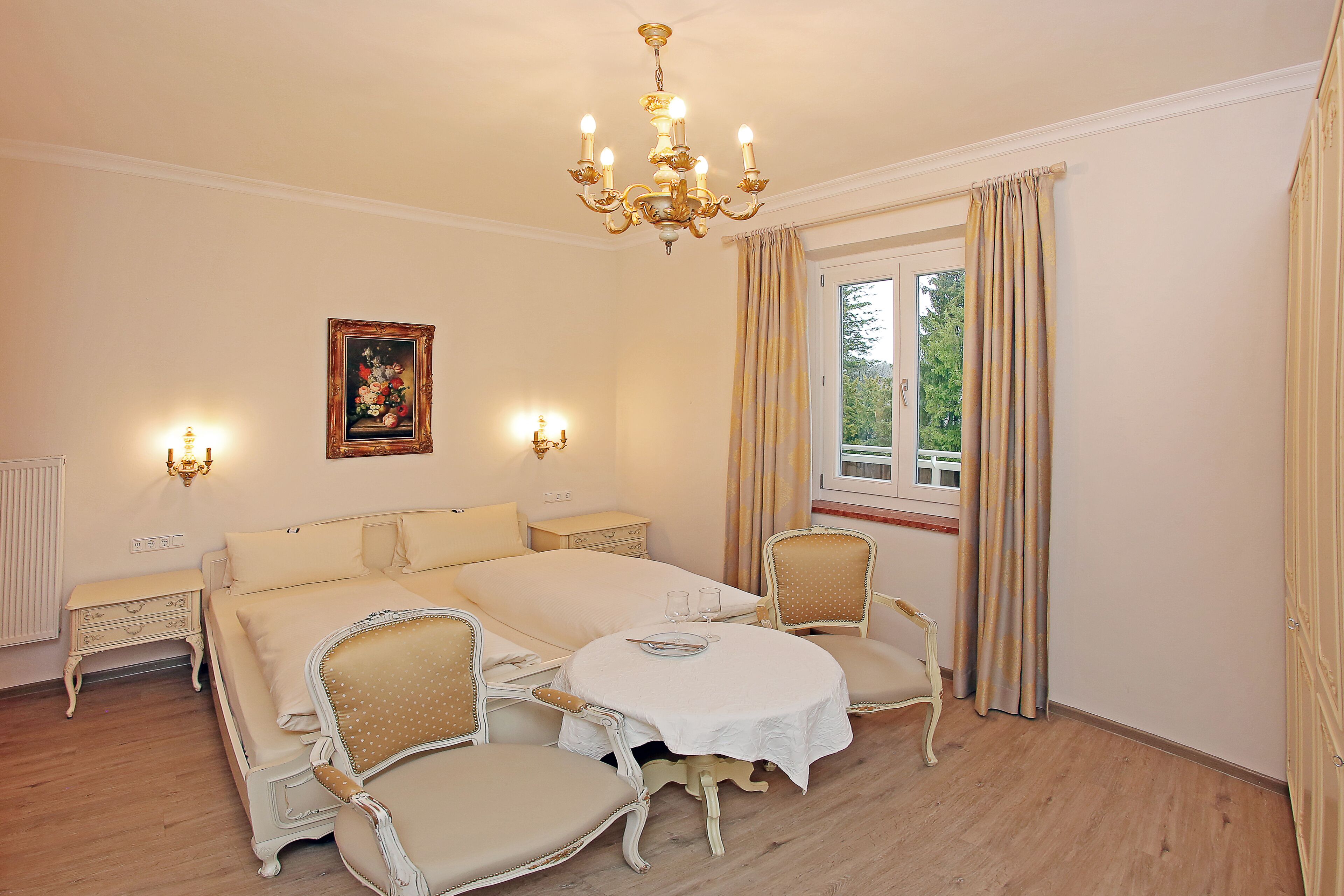 Un cuarto de hotel con una cama grande, dos mesitas de noche, un candelabro, una ventana con cortinas y un cuadro en la pared.