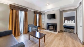 Una habitación de hotel moderna con sofá, una mesita, televisor montado en la pared, chimenea y cama con vistas a través de una puerta abierta.