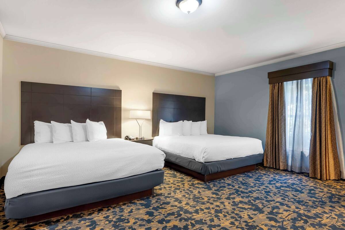 Habitación de hotel con dos camas, un cabecero grande, una mesita de noche, una lámpara y un ventanal con cortinas.