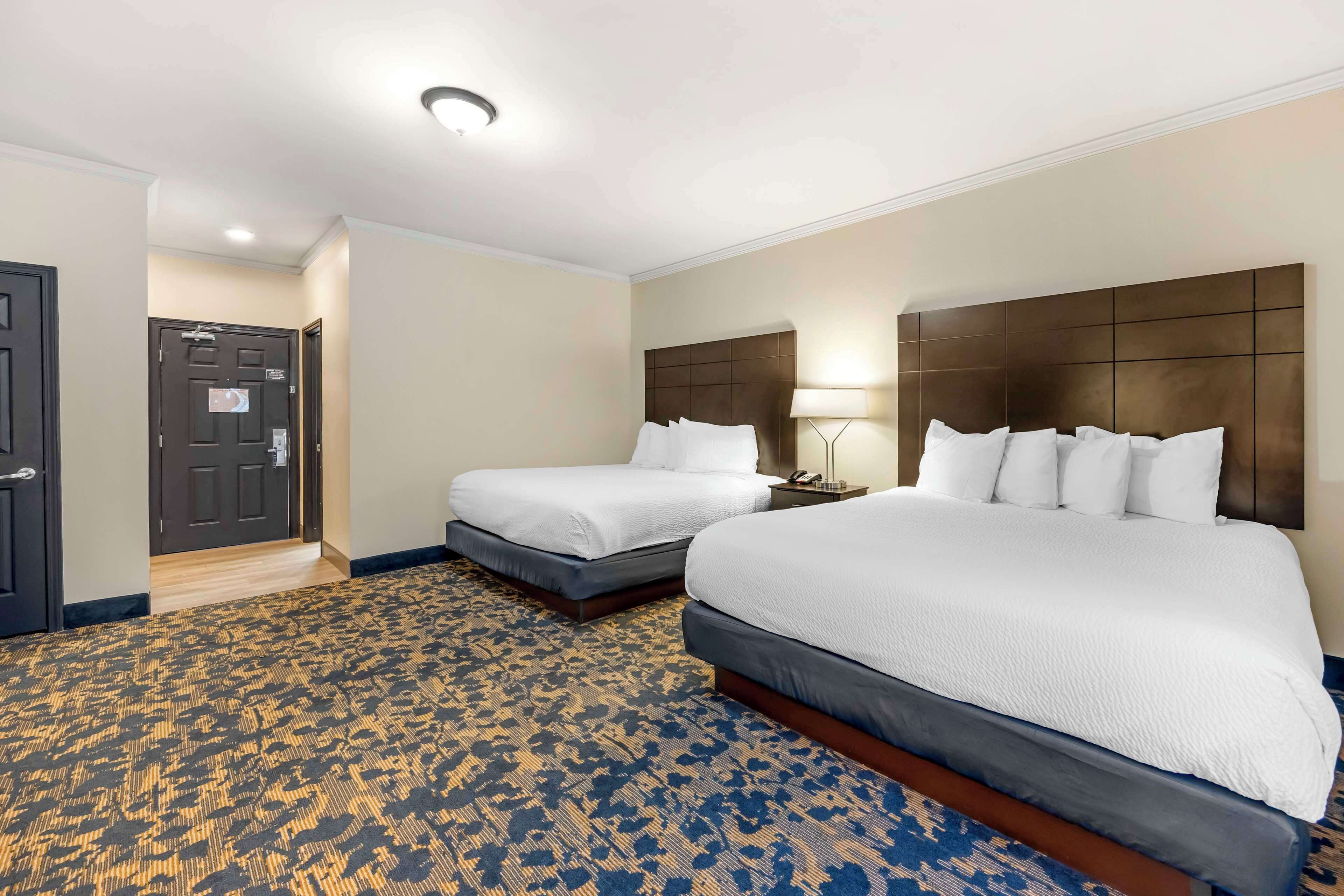 Quarto de hotel com duas camas, um tapete estampado, cabeceira escura e uma porta com uma placa.