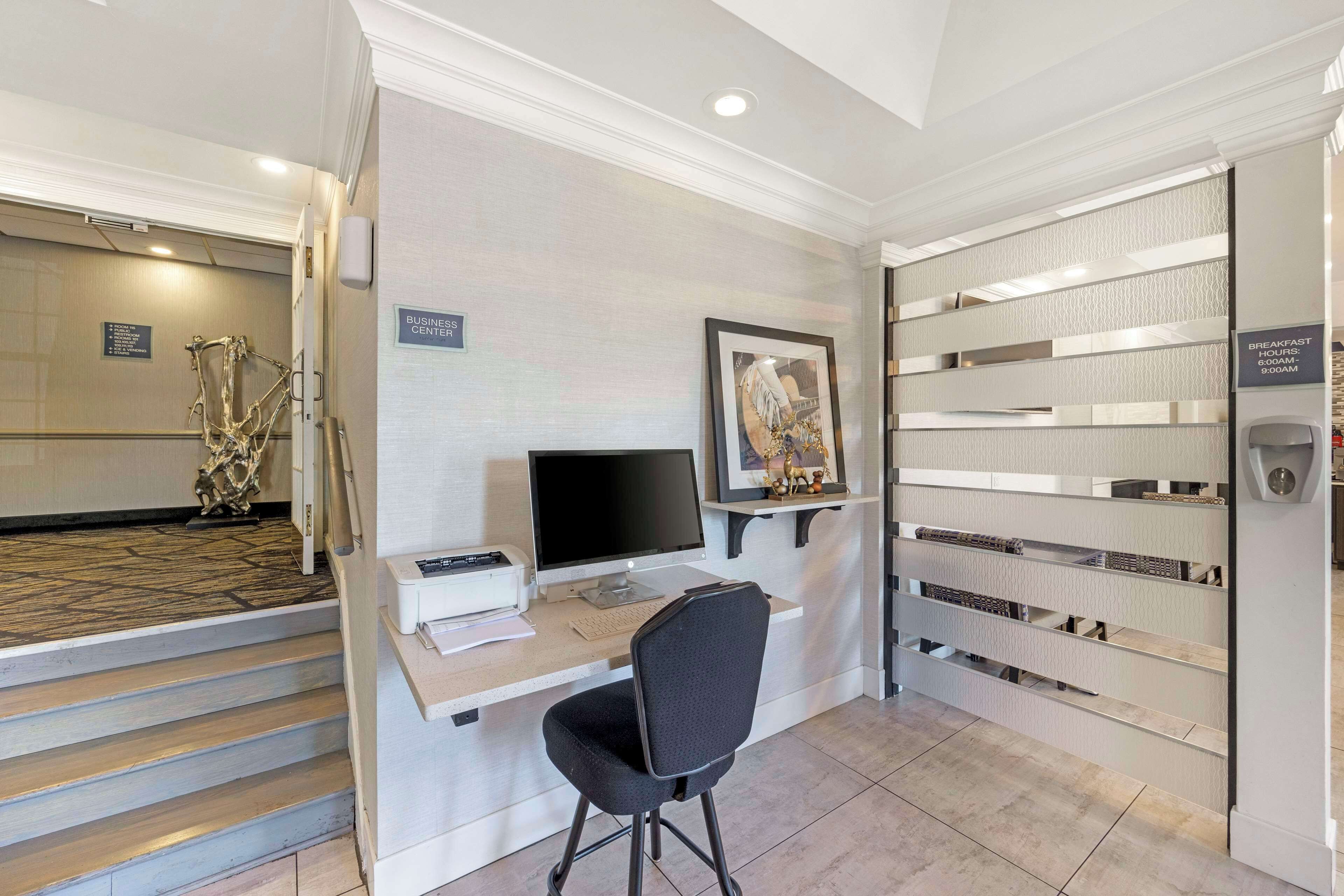 A reception desk with a computer, a chair, and a framed picture on the wall. There is a sign indicating the business center and a sign for breakfast hours.