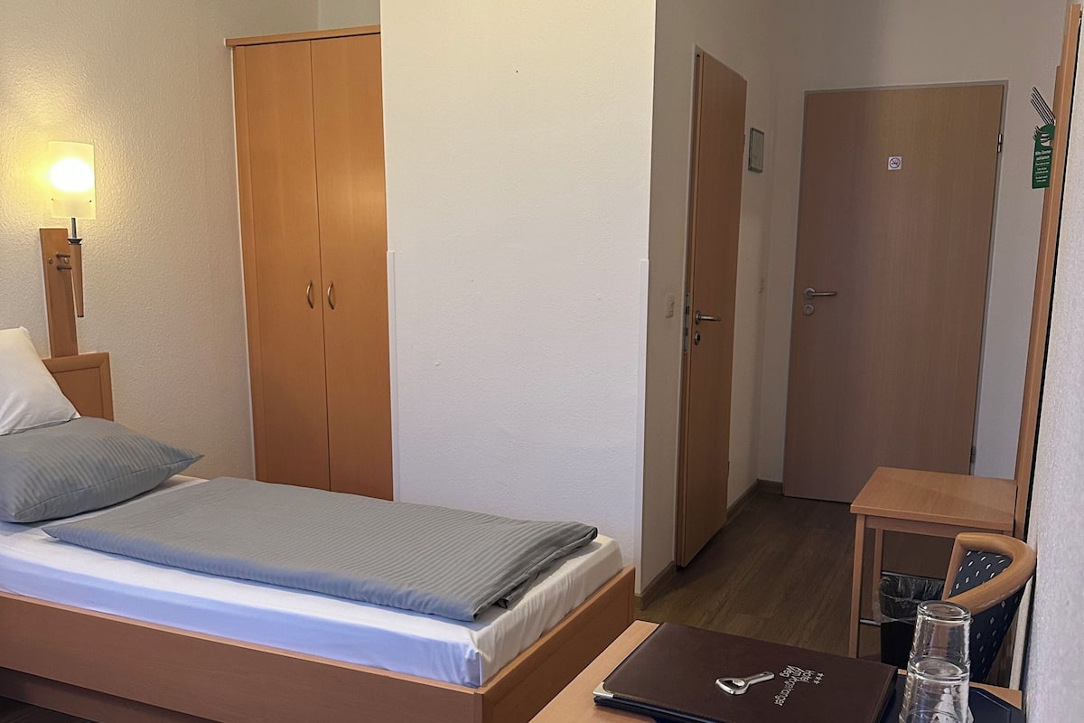 A hotel room with a bed, a desk with a laptop, a chair, and a small table with a lamp.