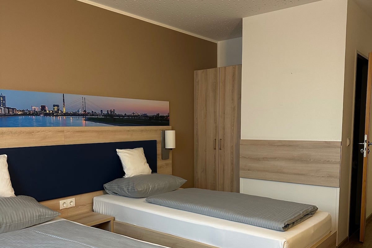 A hotel room with two beds, a wooden wardrobe, a painting of a cityscape, and a desk with a notebook and pen.