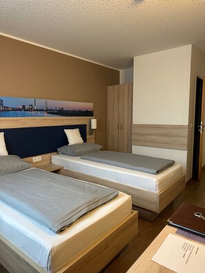 A hotel room with two beds, a wooden wardrobe, a painting of a cityscape, and a desk with a notebook and pen. - Hotel Am Vogelsanger Weg (Düsseldorf)