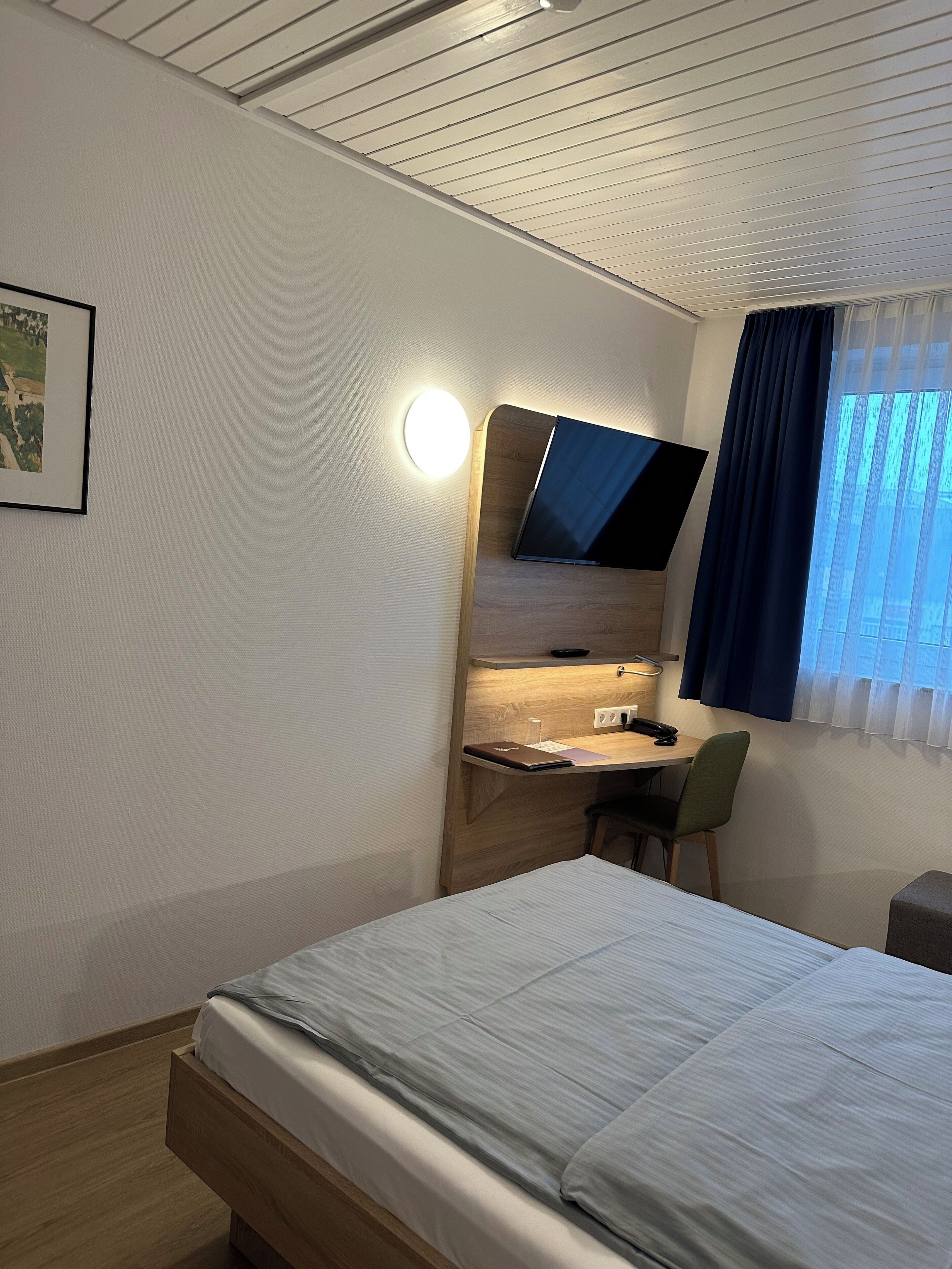 A hotel room with a bed, a desk with a chair, a flat-screen TV, a bedside table, and a window with curtains.