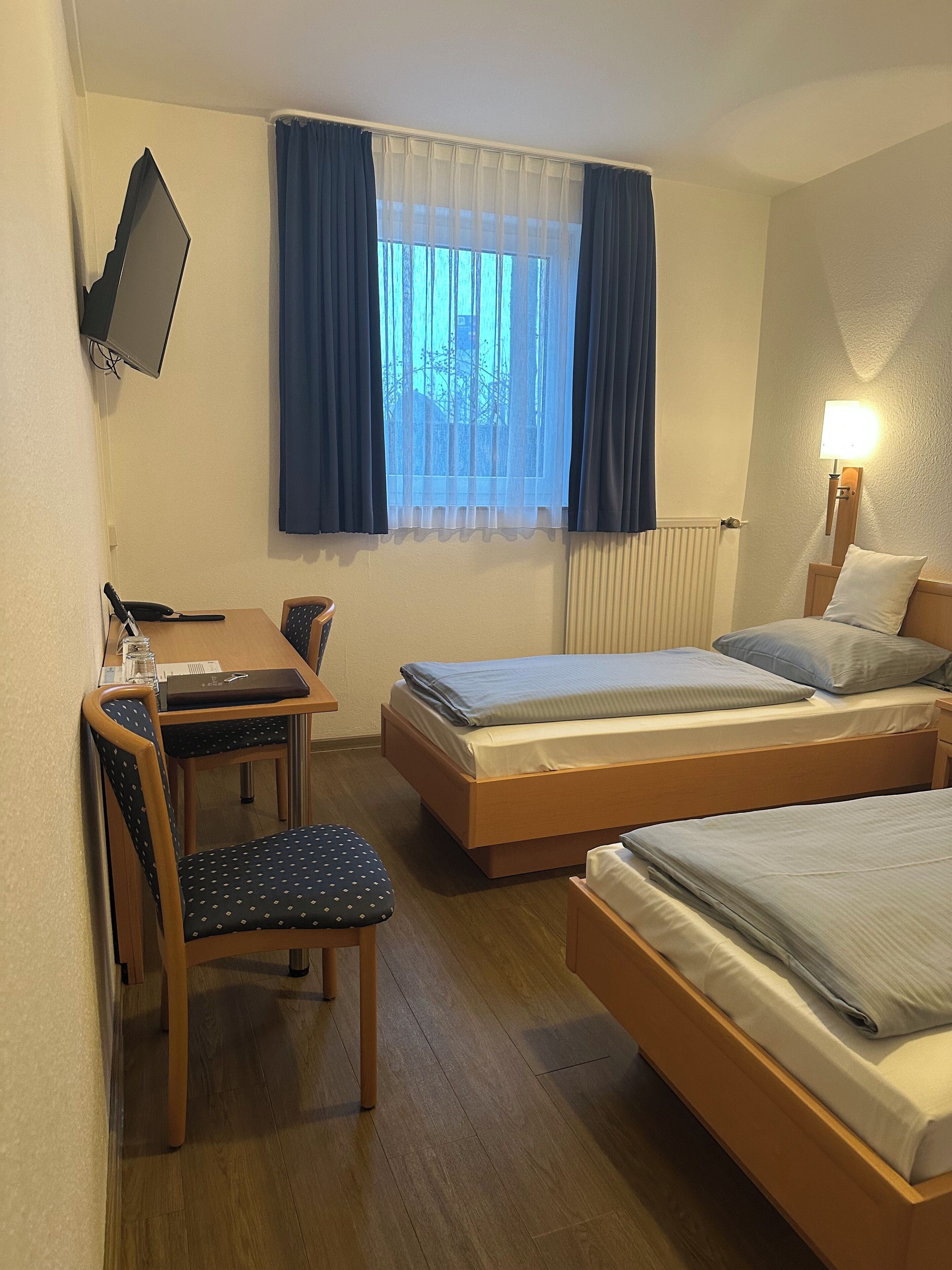A hotel room with two single beds, a desk with a chair, a TV mounted on the wall, and a window with curtains.