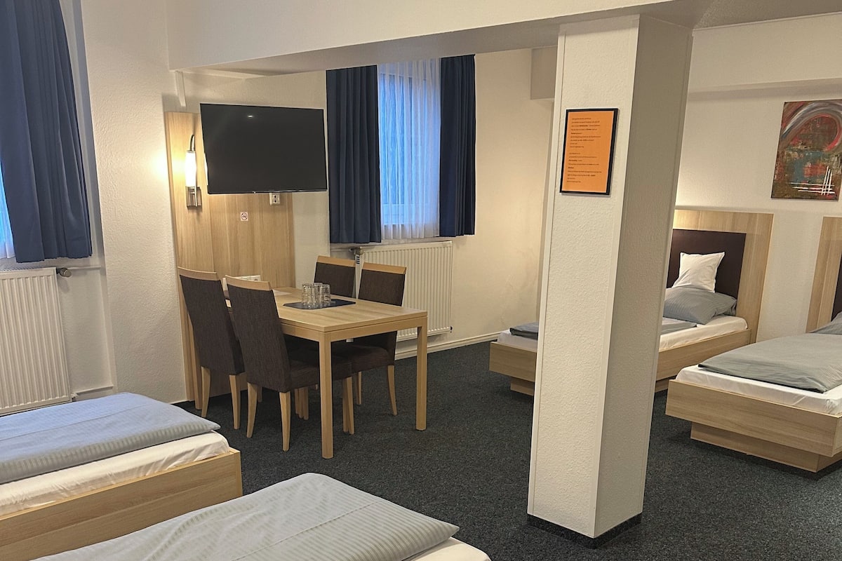A hotel room with three single beds, a wooden desk, a chair, a flat-screen TV, and a window with curtains.