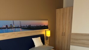 Quarto de hotel com duas camas de solteiro, cabeceira de madeira, mesa de cabeceira, luminária na parede e vista para a cidade.