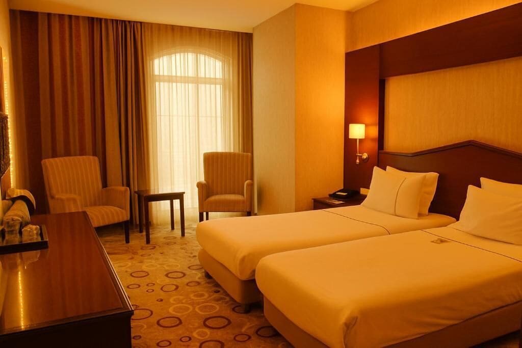 A hotel room with two beds, a desk, a chair, and a lamp.